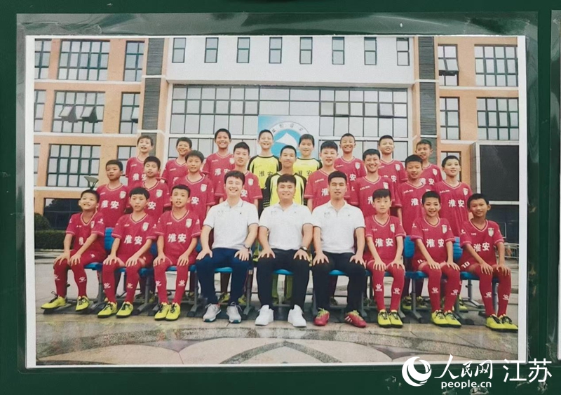 李世恩（上排左四）在淮安工业园区实验学校05-06组足球队的合影张贴在教练室墙上，照片中多名球员正效力于“苏超”淮安队。 人民网 鞠峰摄