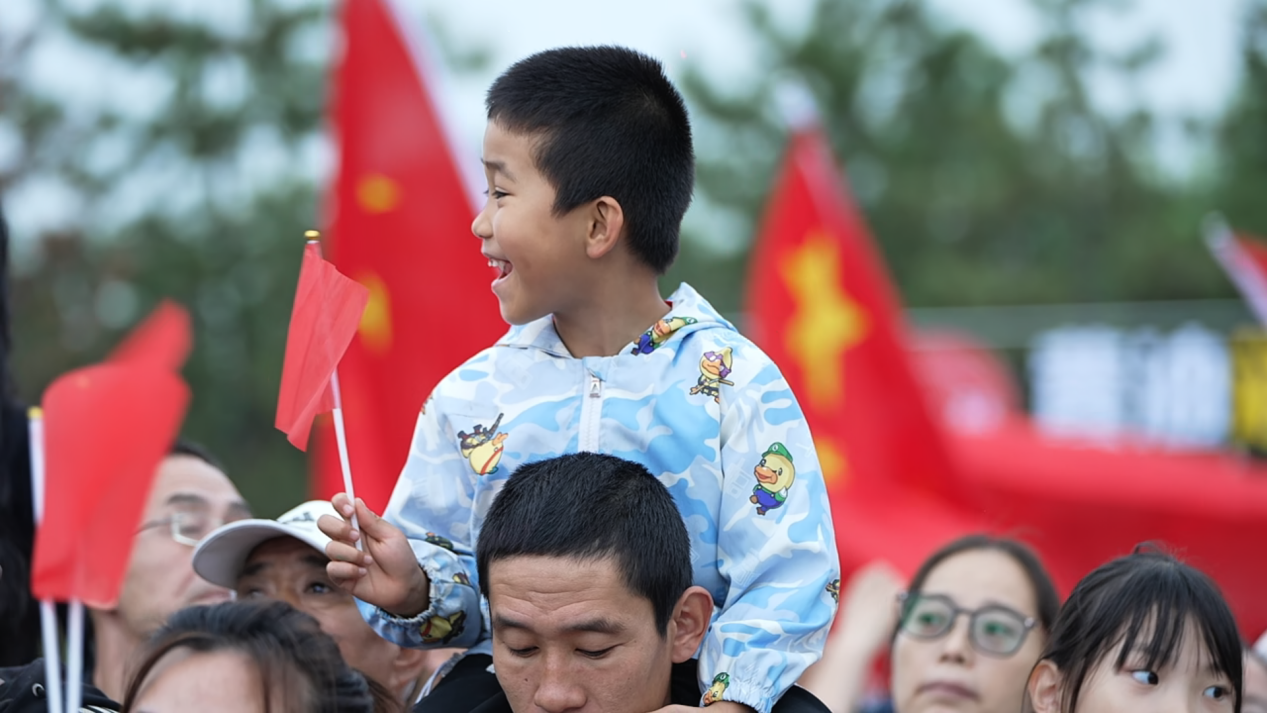 在百狮园南川古渡水路巡游表演区，一名孩童坐在家长肩头，手握小红旗，兴高采烈地观赏展演。贾玉龙摄